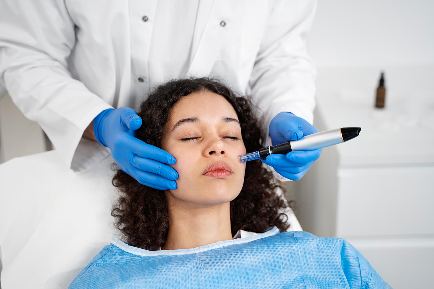 Manso con unos guantes azules sujetan las cara de una chica con los ojos cerrados. Una de las manos sjeta un boli de dermapen