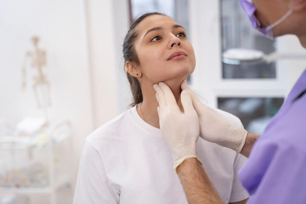 Médico inspeccionando el cuello de una chica. El médico lleva unos guantes blancos y un uniforme lila. No se le ve la cara. La chica tiene los ojos marrones y lleva una camiseta blanca