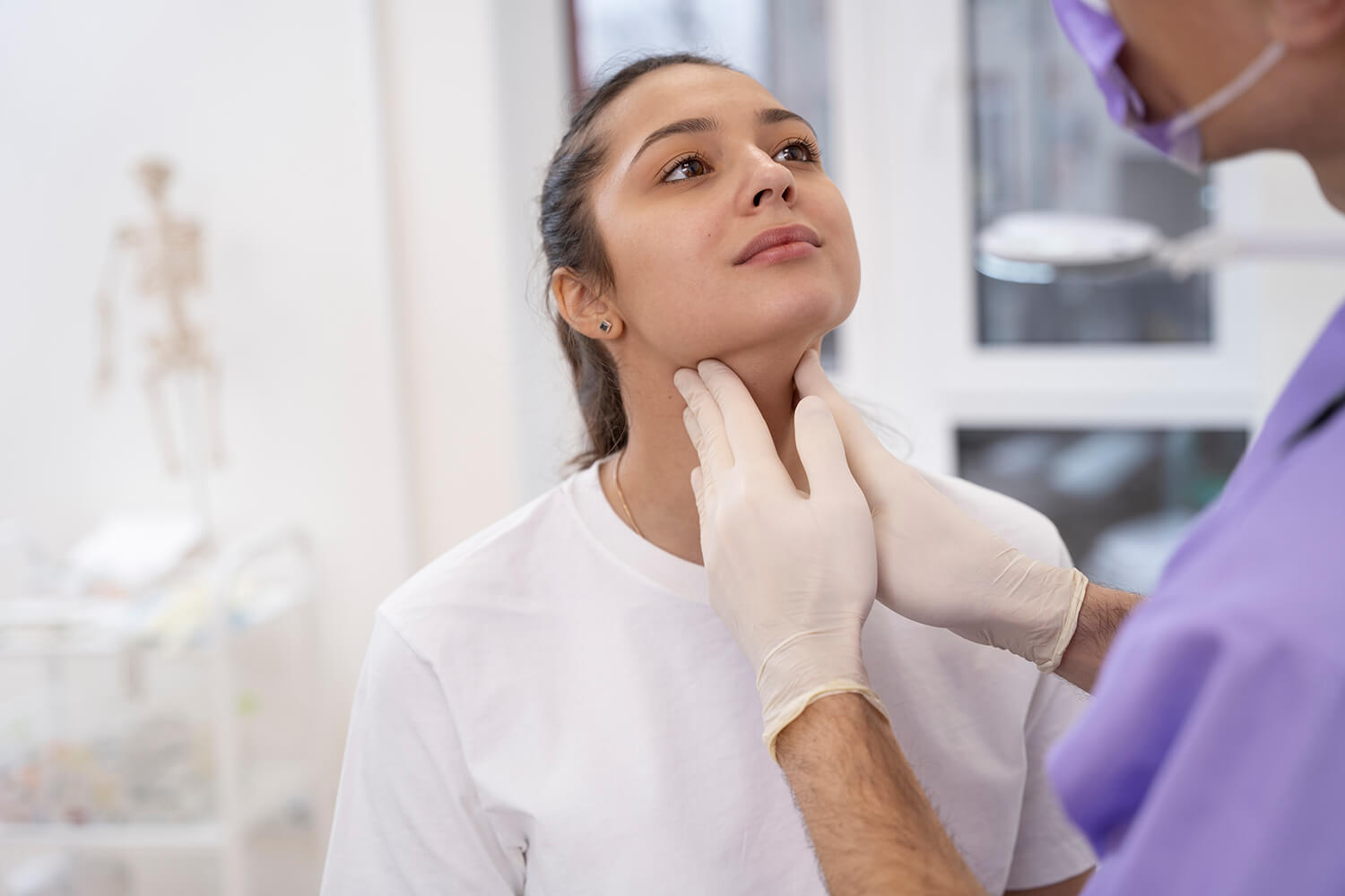 Médico inspeccionando el cuello de una chica. El médico lleva unos guantes blancos y un uniforme lila. No se le ve la cara. La chica tiene los ojos marrones y lleva una camiseta blanca