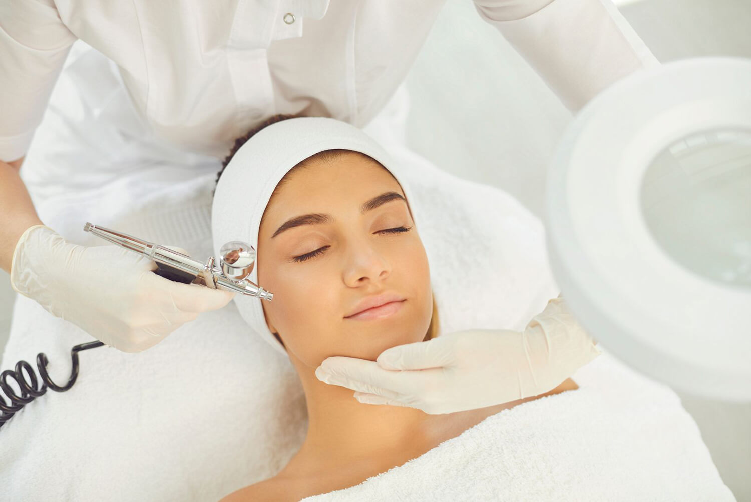 Manos con guantes blancos sujetando una pistola de oxigenoterapia. En el medio hay una chica con los ojos cerrados, esperando a que le hagan el tratamiento